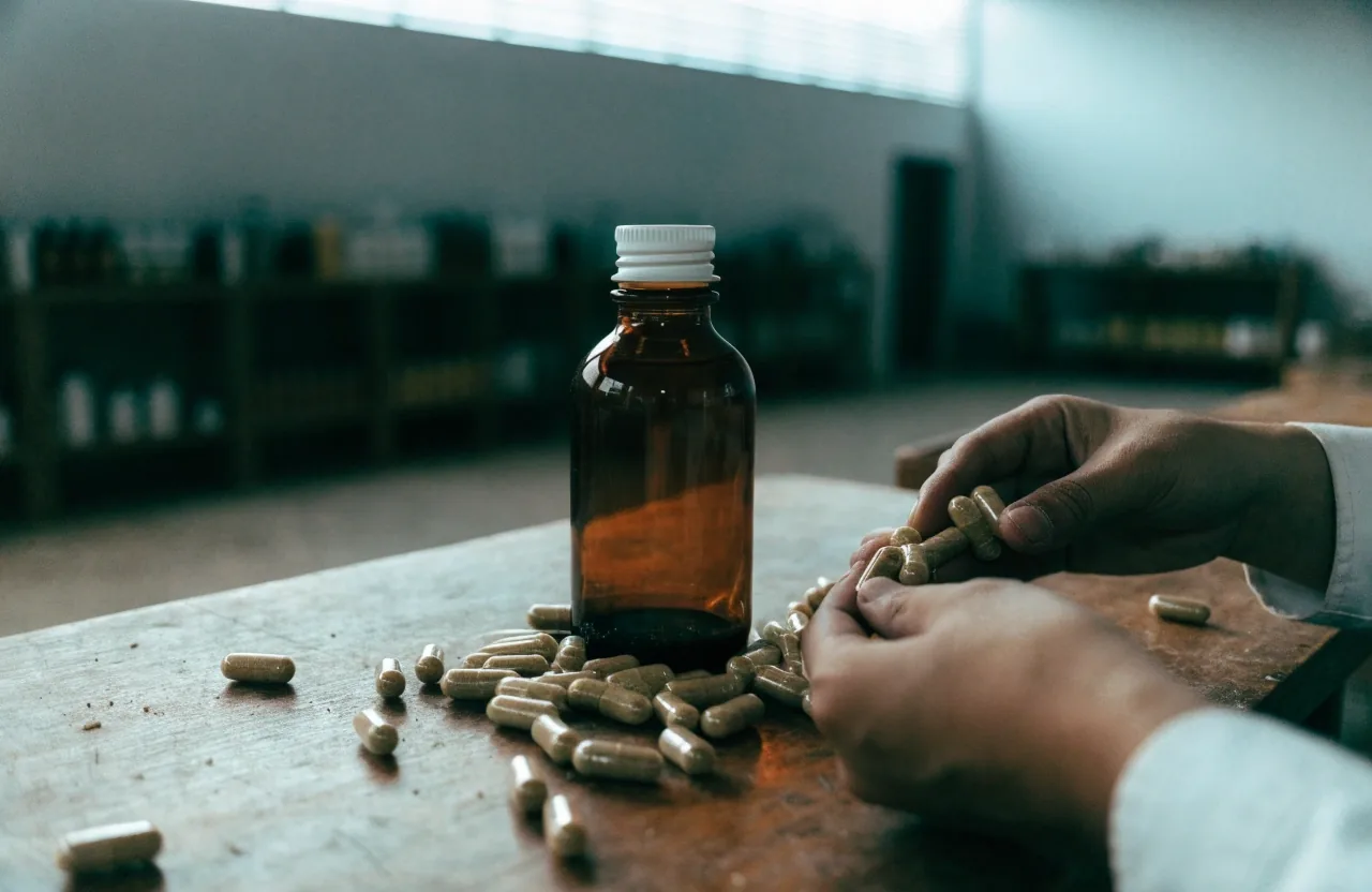 A blank unlabeled bottle of natural capsules standing on a wooden workbench in the VitalitasIndo workshop, loose capsules resting on the wood